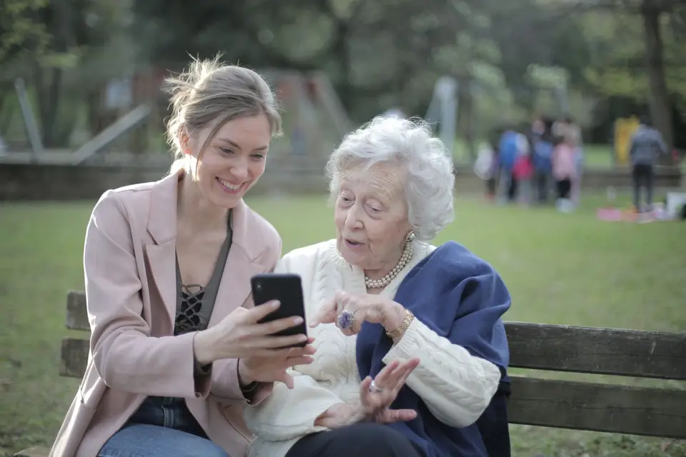 An adult daughter and her mother sit on a bench outdoors, looking at a phone together.